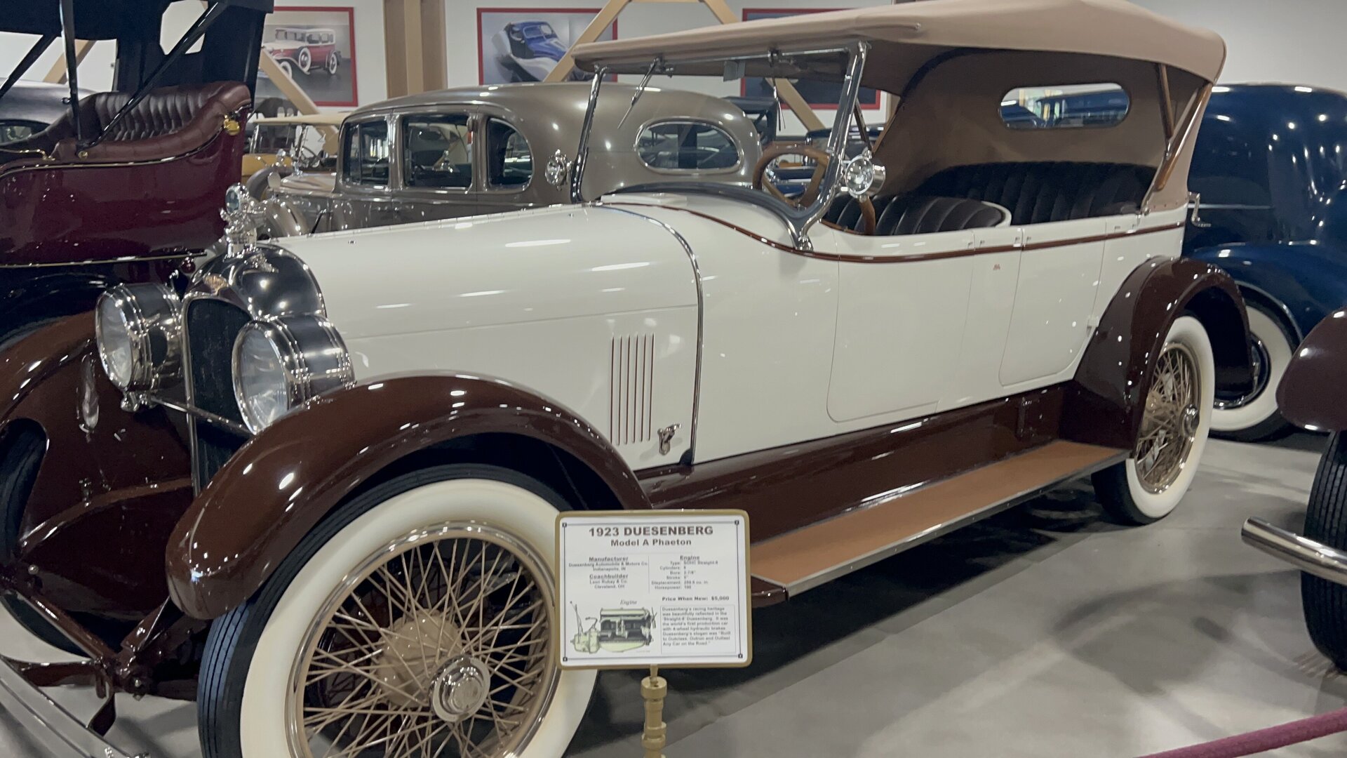 A 1923 Duesenberg Model A Phaeton with a white body, brown fenders, and a tan soft top, parked on a patterned museum floor.