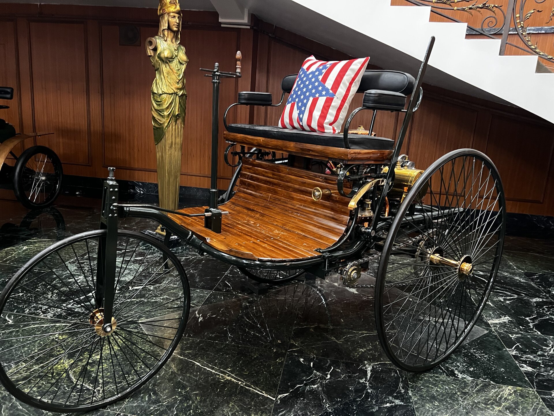 Overhead view of the 1886 Benz Patent-Motorwagen showing the exposed brass horizontal single-cylinder engine, flat cast-iron flywheel, belt-and-chain drive to the rear axle, tufted leather bench, and wooden slat floor.