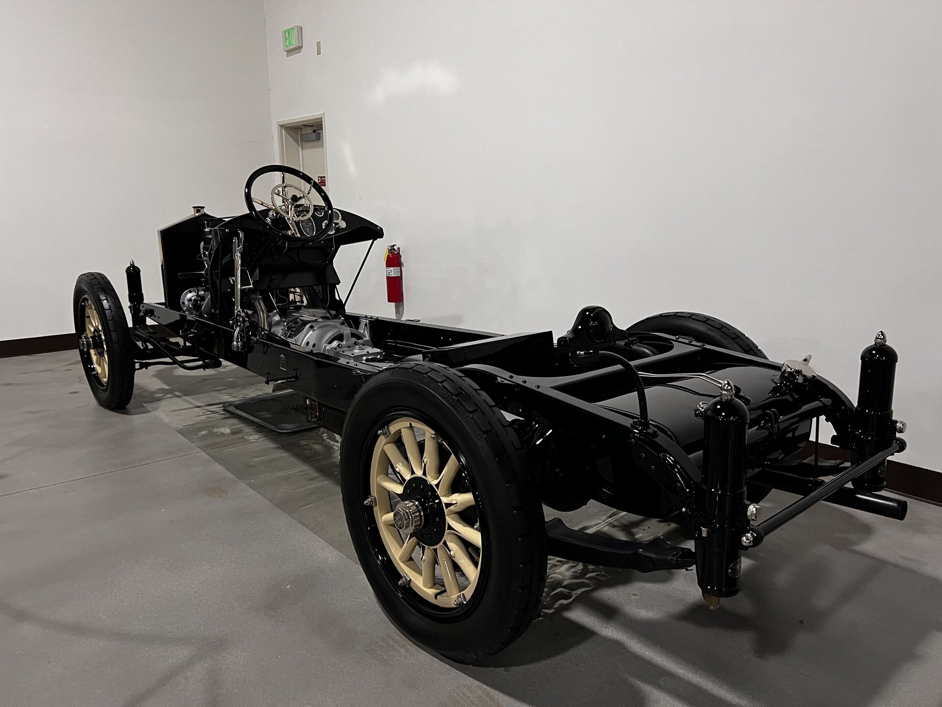 A black 1917 Owen Magnetic Model 60 display chassis at the Nethercutt Museum, viewed from the rear three-quarter angle, showing the exposed magnetic transmission housing where a gearbox would normally sit, and cream wood-spoke wheels.